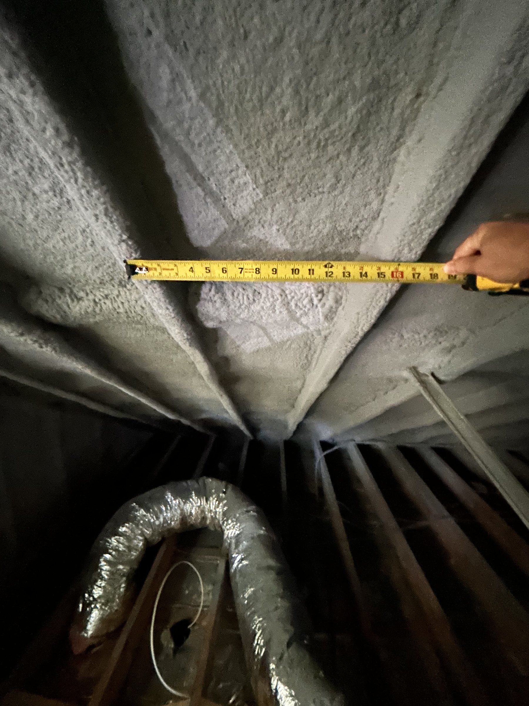 Attic rafters of a 1951 cape covered in spray foam insulation with a tape measure held across them